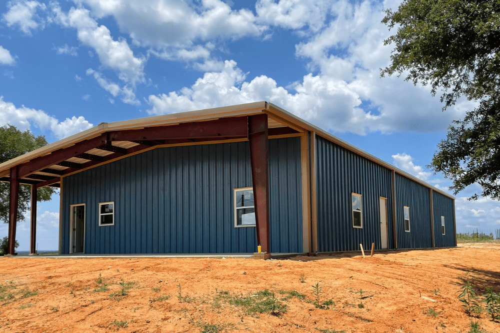 Metal building on a red dirt lot, featuring blue vertical siding, a covered entry, and multiple windows and doors under a partly cloudy sky. Metal Roofing and Metal Building Services In Lacombe, LA, At ADCO Metals.
