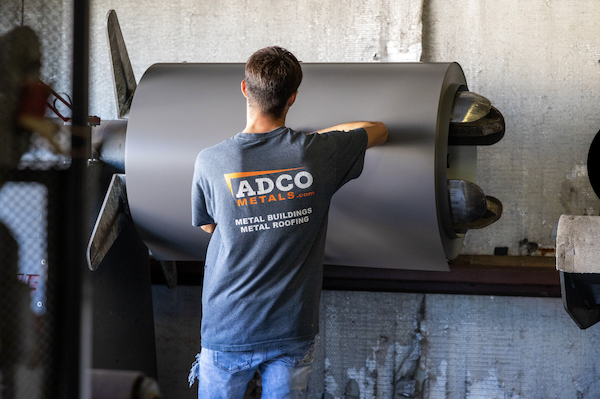 A man rolls out sheet metal in answer to the question is a metal roof cheaper than shingles